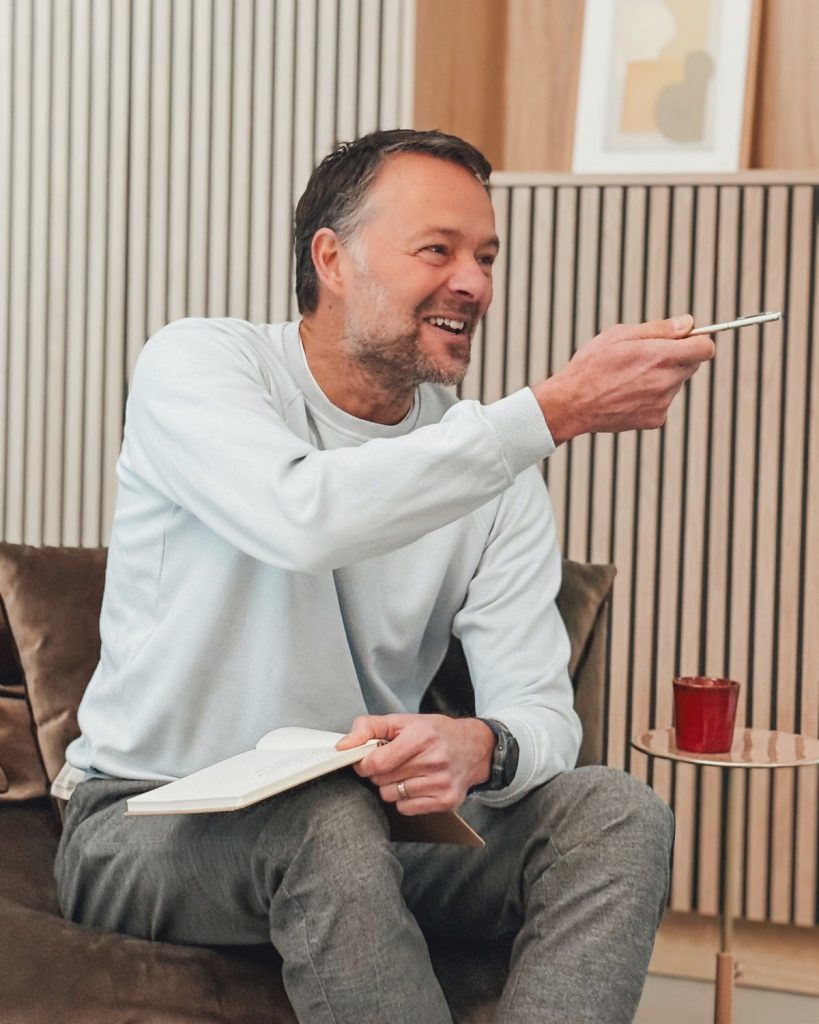 Ivo Hoogland zit lachend op een bank met een notitieboek en wijst met zijn pen